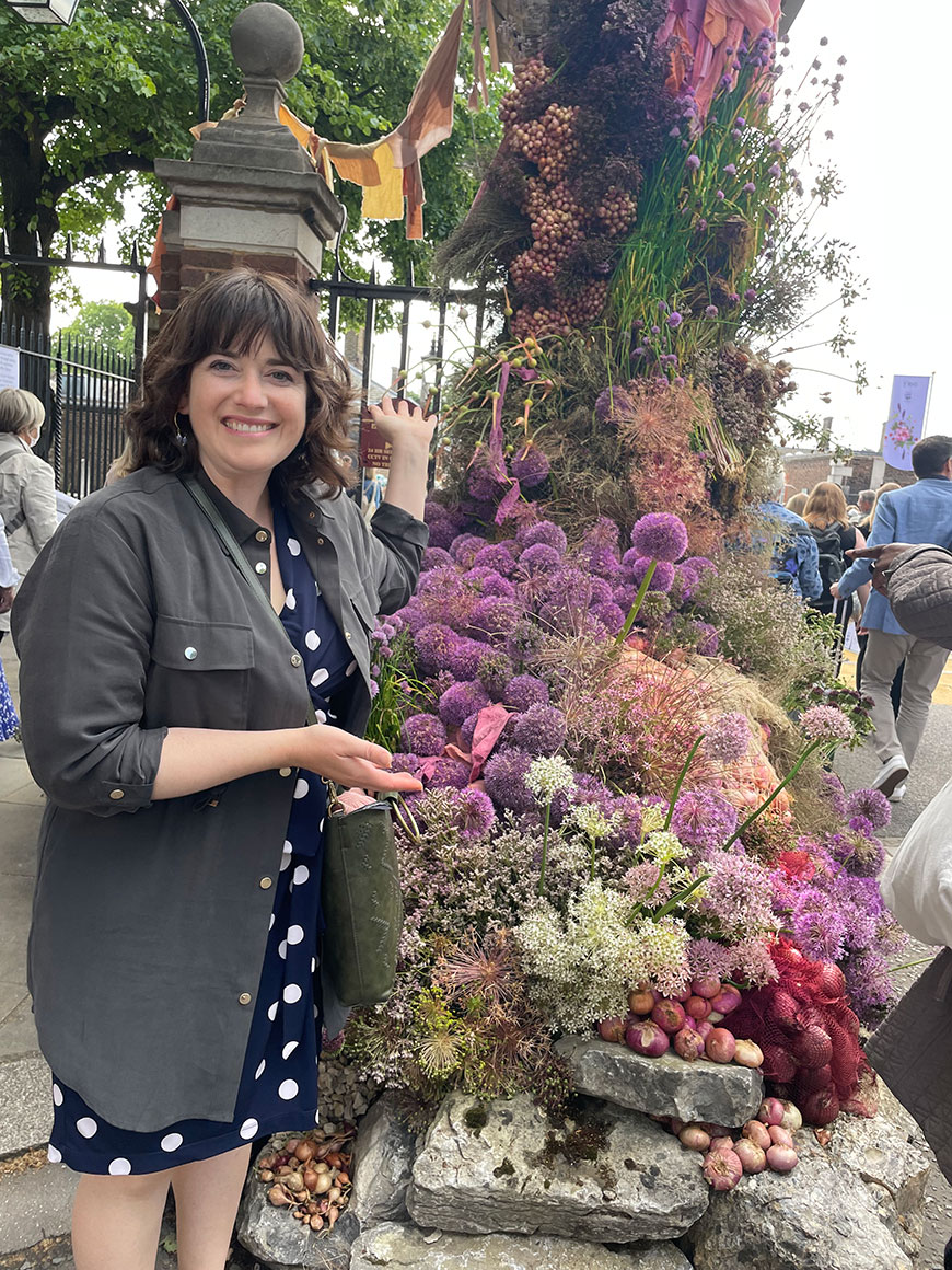 A smiling woman wears polka dots and stands excitedly next to a dried flower arch of Alliums at the entrance to the Chelsea Flower and Garden Show. 