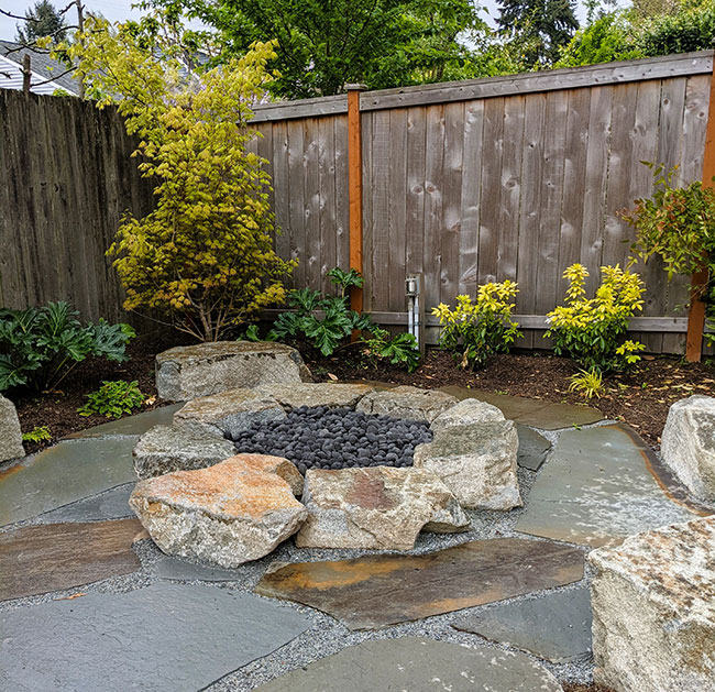 A circular firepit constructed of large stones sits in the center of a flagstone seating area.