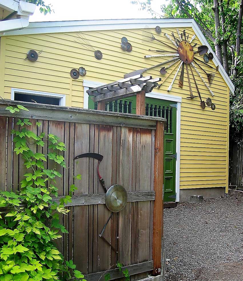 Repurposed rusted saw blades and garden tools form a ‘galaxy’ above a bright green door on a yellow garage wall.