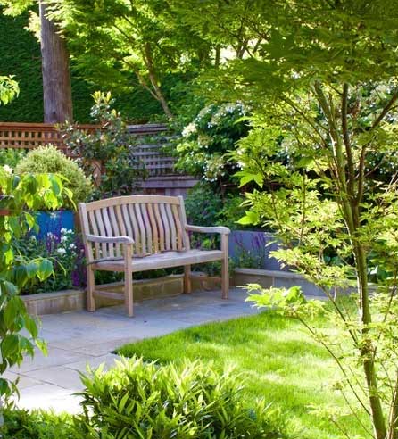 A wooden garden bench sits on a bluestone paver path amidst various shrubs and perennials.