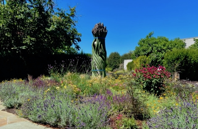 Aphrodite Asparagus, a statue of a hidden goddess peering out from a giant asparagus bundle, is surrounded by a rich palette of perennials.