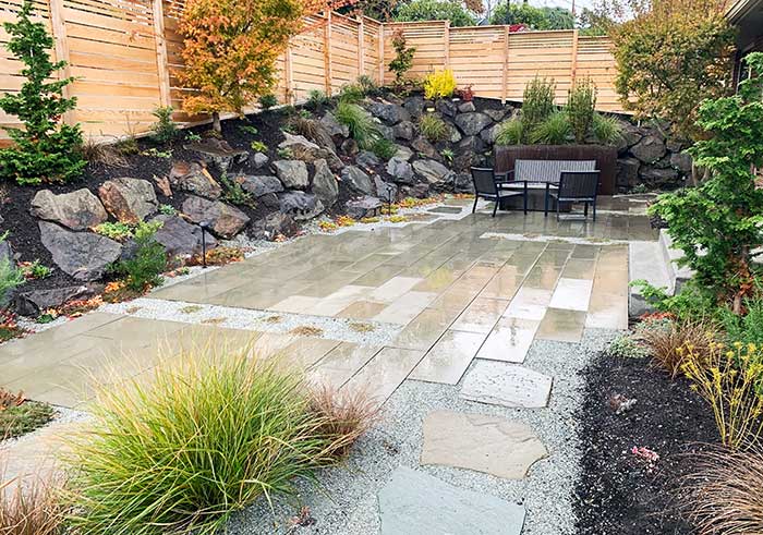 Paver patio with rock wall on the side and various plantings around the patio.