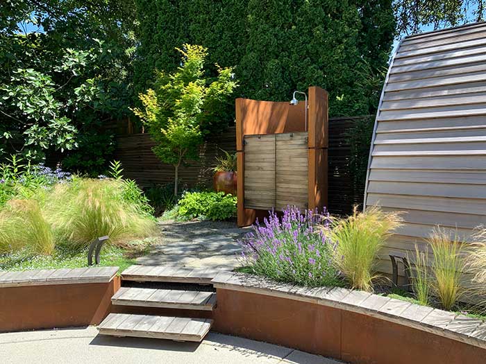 Steel retaining wall with cedar seating around a concrete patio, steps leading to an outdoor shower across from a cedar sauna. Grasses and lavender are planted in the beds by the steps.