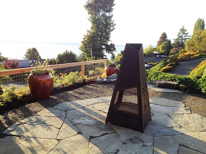 Metal firepit on a flagstone patio that overlooks the Puget Sound. Bright red containers and plants brighten up a low fence made of hog wire and cedar. 